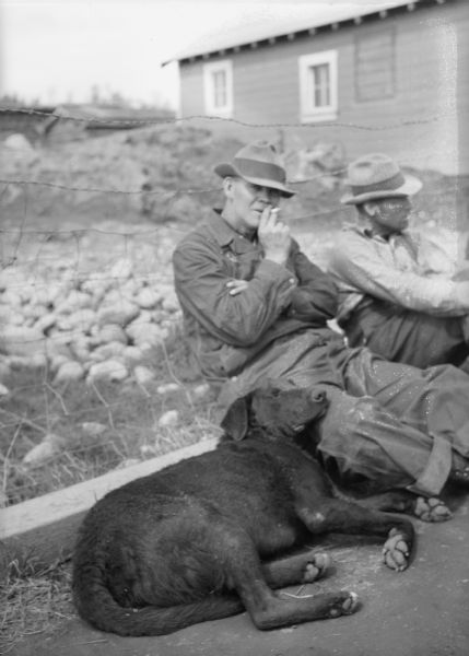 Two men are sitting along a fence, and a large dog is lying near them. There is a building in the background. Caption on back of print reads: "May/June 1035 in the Matanuska. At times, there isn't much to do but wait."