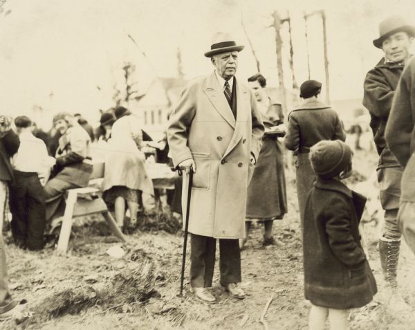 Caption on back reads: "Gov. Parks. Colony day 1936. Ruth Dearmond behind him (right)." There is a child standing in the foreground, and many people are gathered in the background.