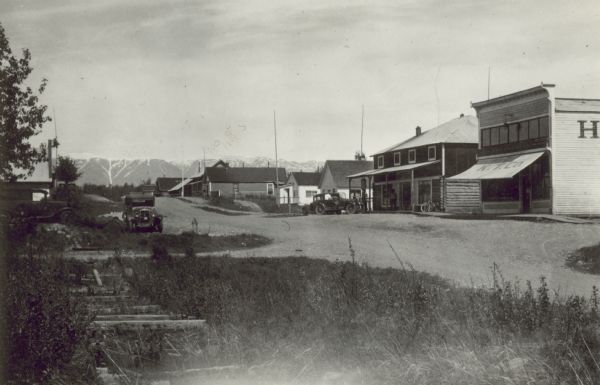 View towards Palmer, with commercial buildings, including the Wasilla Road House, and the K.T. Co. Automobiles are parked along the edge of the street, and snow-capped mountains are in the distance. Caption on back reads: "It didn't take Palmer, at first a one-building railroad town, long to develop its own main street."