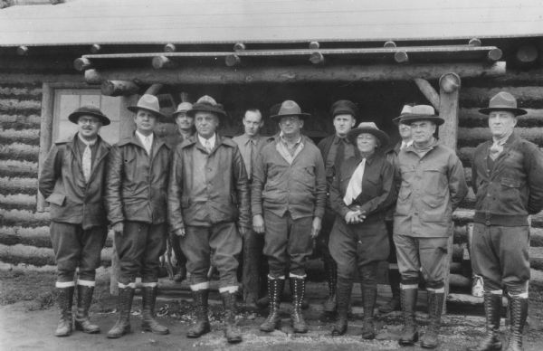 Outdoor group portrait of men and women standing in front of a log building.