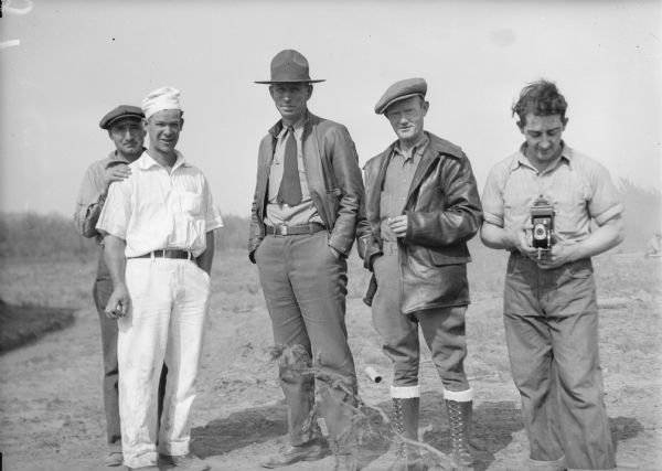 Original caption reads: "Ben Jordan (tallest), steward at construction division mess, and the boys who feed me." A man standing on the right is holding a camera at his waist.