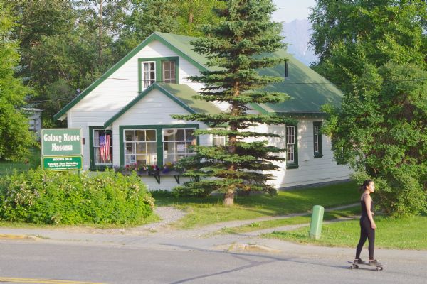 View from road towards the Colony House Museum. A sign outside the museum reads: "This house was constructed in 1935 as the farm home of a Matanuska Valley Colonist family. It was relocated in July 1995 from Farm Tract No. 94 to this permanent site within Palmer's Historic District. Working together to preserve our local history, many residents and supporters of this Museum project have brought about the restoration of these buildings. We invite you to experience the history of Palmer, Alaska 1935-1945 when you visit the Colony House Museum. The Palmer Historical Society."