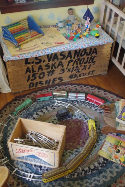 A child's bedroom in Colony House Museum. A toy railroad on a braided rug is on the floor. Dolls, toy blocks, and a coloring book are also on display. A shipping crate from the 1935 journey from the Midwest to Alaska serves as a toy chest.