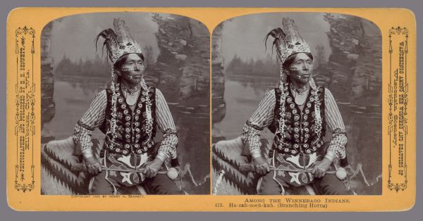 Waist-up studio portrait in front of a painted backdrop of Ho-Chunk man Branching Horns in full traditional dress. Caption at bottom: "Among the Winnebago Indians. 413. Ha-zah-zoch-kah. (Branching Horns)." Printed below the left side stereograph: "Copyright 1905 by Henry H. Bennett." Text at right: "Wanderings Among the Wonders and Beauties of Wisconsin Scenery."