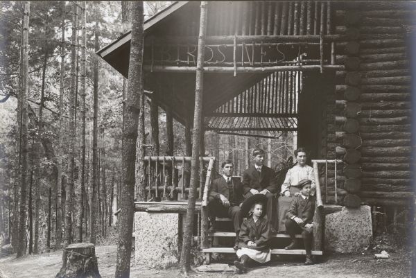 A group of men, women, and children are sitting on the porch steps of a log cabin in the woods. The words "Birds Nest," fashioned out of bent sticks, are above the steps along the side of the porch roof. 