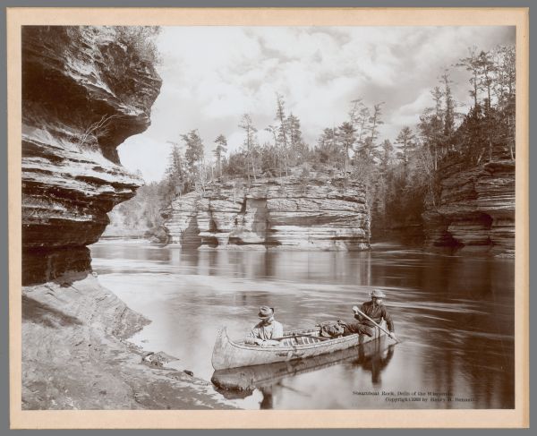 View from shoreline towards two men in a canoe on the Wisconsin River in the foreground. There is a rock formation along the opposite shoreline.