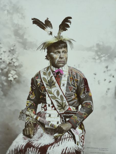 Waist-up studio portrait in front of a painted backdrop of a Ho-Chunk man, Chach-scheb-nee-nick-ah (Young Eagle) looking off to the right. This photograph has hand-applied color on the intricate beadwork on the subjects clothing and accessories.