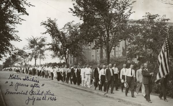 Kohler strikers marching along a sidewalk. Handwritten on the photograph: "Kohler Strikers '2 years after' Memorial Picket Line, July 26th, 1936."
