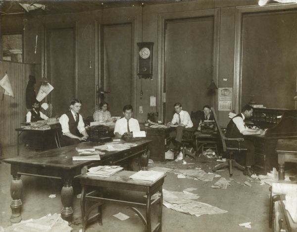 Staff of Chicago Daily Socialist working at desks in a large, open room. Caption reads: "Staff of Chicago Daily Socialist. L to R.: A.M. Simons, W. Beyer(?) May Wood-Simions, John Carroll, remainder unidentified." There is a 1909 calendar in the background.