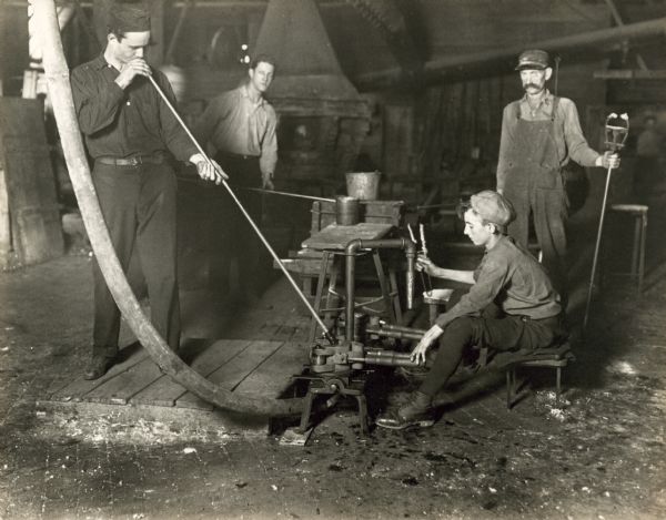 Group of men glassblowing. The boy at right is working the mold for the glassblower.