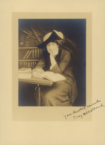 Studio portrait of suffragist Inez Milholland sitting with her chin in her hand at a table with an open book. Signed at bottom right: "Your devoted comrade Inez Milholland."