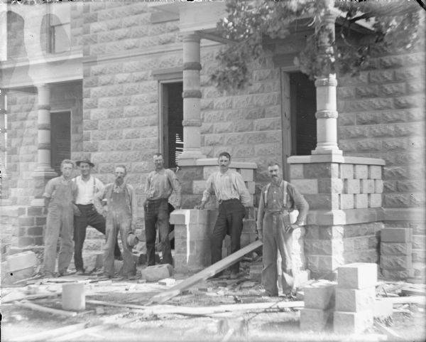 Six men in work clothes posing near a porch of a house under construction. Hollow core concrete blocks, most likely manufactured on site with local sand and aggregate, form the walls of the house and are stacked on the ground. Round concrete blocks have been used for the porch columns. A note on the negative envelope states: "Carpenters Mr. Wing and Mr. Day."
