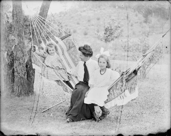 A young woman is sitting between two girls in a hammock. The girl at right has a large bow in her hair and is wearing a necklace. 