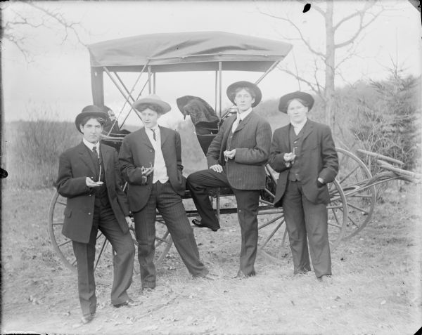 Four women dressed as men posing standing in front of a buggy. They are wearing hats and suits with vests and ties. Each woman is holding a pocket watch. A note on the negative envelope identifies: "Agusta, Leta [von der Sump], Marie and Emma Dressed Up."  