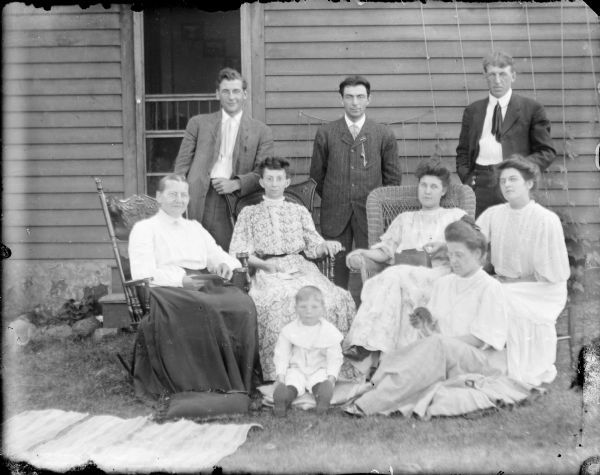 A group including members of the Von der Sump family posing near a house. Three men stand in the rear and include, from left, Fred von der Sump, Ellis Owens and Will von der Sump.  Seated from left are Bertha von der Sump, Mrs. _____ Wallace, Leta von der Sump and an unidentified young woman.  An unidentified little boy sits on the ground; Hattie von der Sump Owens is also seated on the ground and is looking at a booklet. There is a rag rug and pillow on the ground. Behind the group, wooden steps lead to a screen door.
