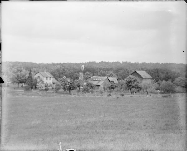 A view across a field towards a farmstead, including a wood frame two-story house, barns and sheds, and a windmill. There is a fence at the edge of the field and a low wooded ridge in the background.   