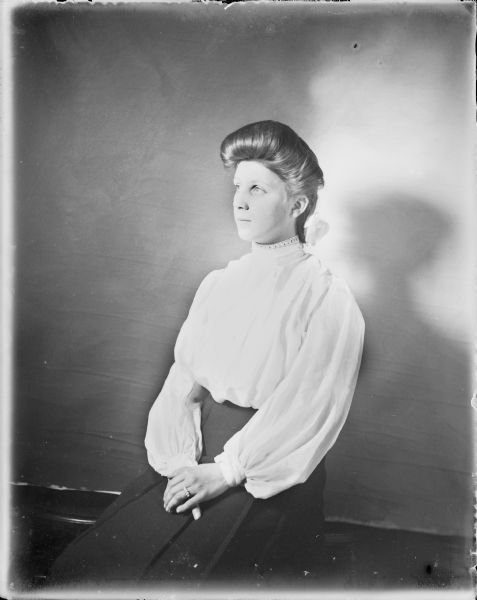 A seated, three-quarter length portrait of a young woman wearing a light-colored blouse and dark skirt. She has a ribbon in her hair, and a ring on her finger set with multiple oval stones.