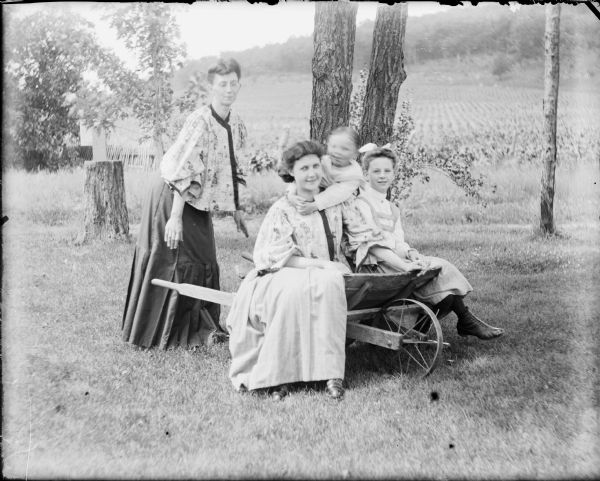 A woman, identified as Mrs. Wallace, is bending forward as if to grasp the handles of a wooden wheelbarrow. Leta von der Sump, left, and a girl identified only as Laurine, right, are sitting on the sides of the wheelbarrow as a little boy, Loyd (blurred from motion), is embracing Leta from behind. Mrs. Wallace and Leta are wearing similar jacket-style blouses. A field planted with a row crop is in the background beyond a fence. 