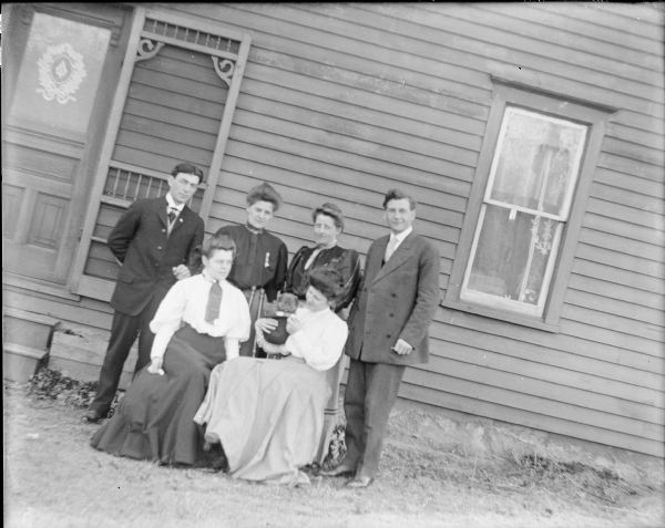 Four women and two men posing near the door of a house. Two of the women are sitting in front of the others, and the woman sitting on the right is holding a puppy in a hat. A handwritten note on the negative envelopes states: "At Hattie's. Mrs. Porter with us." At far right is Fred von der Sump.