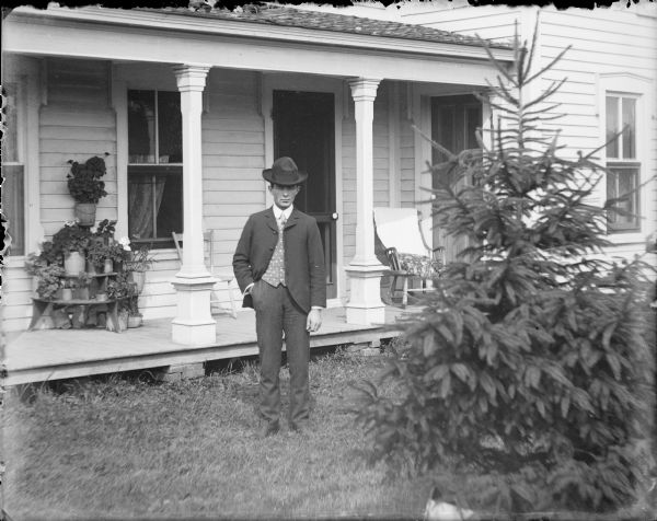 A young man identified as Ellis Owens is standing on the lawn in front of the porch of a wood frame house. He is wearing a suit with a polka dot vest, and a hat. There are chairs on the porch, as well as a wooden stand which is holding potted geraniums and other plants. A small pine tree is in the right foreground.