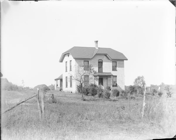 Short cedar shrubs and a small tree are growing in the front yard of a two-story brick house. A second floor door opens onto the flat roof of the front porch. There is a small boy standing at the rear of the house, left, where there is a wood frame extension. There are hay stacks behind the house and fence posts in the foreground.