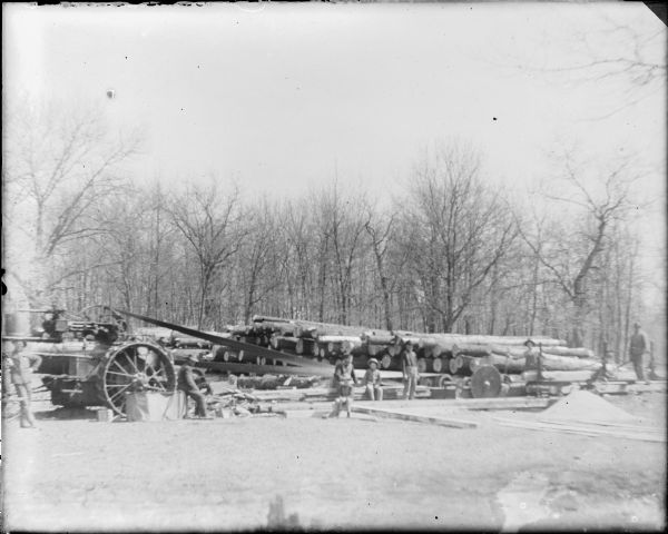 Six men and two boys are posing near a portable saw mill. There is a steam tractor at left, with a long belt driving a large circular saw blade at right, with a pile of sawdust nearby. Logs are stacked in the background. The caption likely refers to the Thomas Kearns farm, also known as Hickory Hill, which was one of John Muir's boyhood homes.  