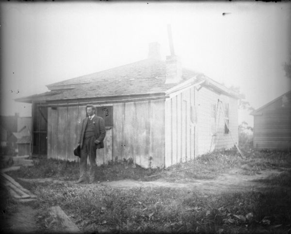 A young man wearing a suit and tie and holding his hat is posing standing in front of a small wood frame house. There appears to be a lean-to addition on the front of the original house. A plank walk leads to the door of the house. A handwritten caption on the negative envelope states: "Fred and the house where he was born in Cambria."