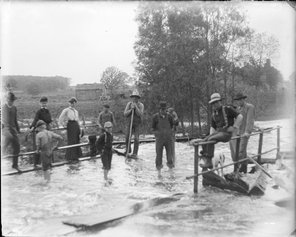 Water from French Creek covers the surface of a bridge at Dates Mill near the Marquette-Columbia County line. The caption on the negative envelope states: "Mae, Leta [Von der Sump] and the boys on the bridge at Dates." Several men, boys, a dog and two women are on the bridge. There is a shed in a field in the background.