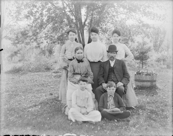 Mary and Thomas Duffy, sitting, are posing outdoors with their five children. Nellie and Thomas are sitting on the ground in front, and daughters (not necessarily in order) Lauretta, Bessie, and Nellie are standing behind them. Flowers are growing in a half barrel on the ground on the right.