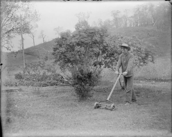 Will von der Sump, wearing work clothes and a hat, is pushing a reel lawn mower in front of a low fence near shrubs. There is a wooded hill in the background.