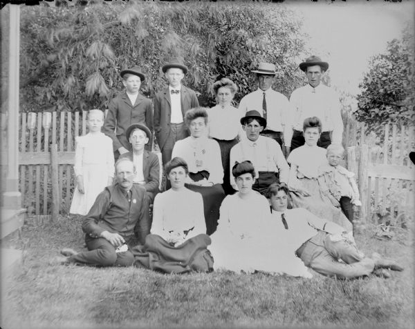 In a photograph captioned "Group of Young Folks at Uncles," well-dressed children and young adults are posing with an older gentleman, who is at front left. Several people in the group are holding small bouquets. Will von der Sump is second from right in the back row; his sister Mae is sitting at far right with a boy leaning against her, and their brother Fred is reclining on the ground, at front right. A corner of a porch is seen at far left, and  a fence is behind the group.