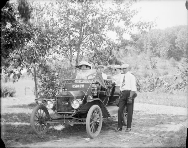 Will von der Sump posing standing beside a 1912 Ford Model T Touring car.  He is wearing dress slacks, a white shirt, necktie and leather driving gauntlets. His sisters Leta, in the front seat, and Mae, in back, are passengers in the car. The young man in the back seat is not identified. A sickle bar mower is standing in the background. A "Browning" pennant is attached to the windshield support. The area in southern Marquette county where the Von der Sump farm was located was identified as Browning because a local post office bore that name.