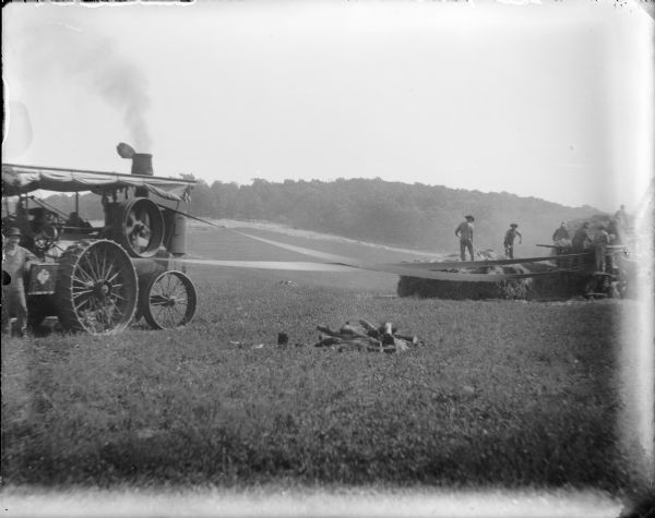 A man at far left, possibly Frank von der Sump, is posing with his elbow resting on a steam tractor. A belt from the tractor is powering a threshing machine at right, where a crew of men are working. There is a small pile of firewood in the foreground.  