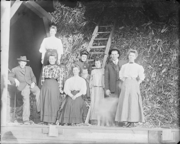 Eight young men and women posing in a barn. They include Will von der Sump, far left, and his brother Fred, second from right. A dog in front of Fred moved during the exposure. Also pictured are sisters Leta, sitting on a ladder holding a cat, and Mae von der Sump, standing next to Leta. The other four women are unidentified.  There is fodder, possibly sorghum, stacked behind the group. 