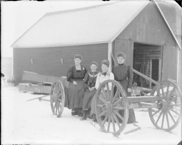 Three women are sitting on the frame of a farm wagon while a fourth woman is standing next to them on the right. The women are well-dressed, but are not wearing coats or gloves. There is snow on the ground and on the roof of the granary behind them. A note written on the negative envelope identifies them (in no stated order) as: "Hattie, Leta, Anabelle and Emma."