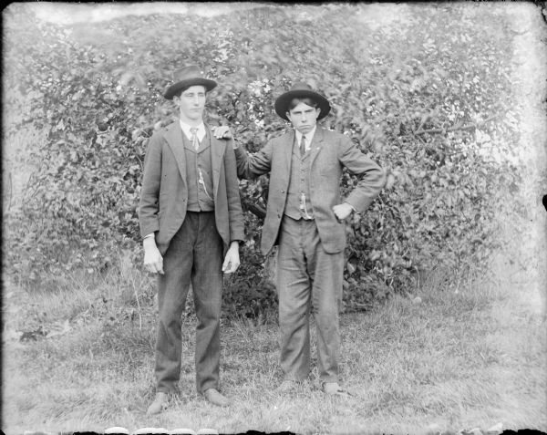 An outdoor, full-length portrait of two young men standing and wearing suits and hats. Alec Robarge is resting his right hand on the shoulder of Henry Hernkind. Robarge was a hired man on the farm owned by Henry's father August.