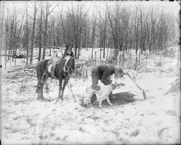A young man identified as Will Koeppe is leaning forward, resting his left hand on a shovel while inspecting a hole in the ground at the edge of a wooded area. There is a dog standing next to him. There is light snow on the ground. A horse with saddle and bridle standing on the left.