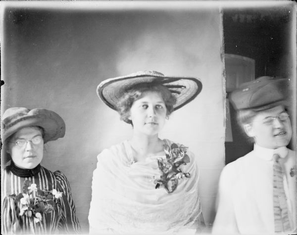 A waist-up portrait of Leta von der Sump, "draped" in a sheer material, and wearing a large hat and corsage. She is flanked by her sisters Mae, left, and Hattie, right. Hattie is wearing a blouse with a collar and necktie. There is a photographer's backdrop behind Mae and Leta.