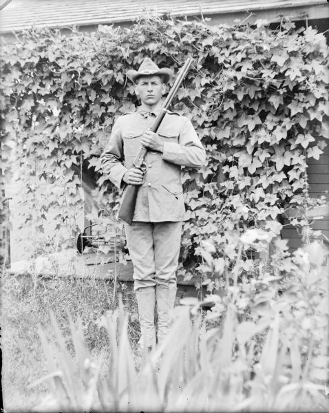 Full-length portrait of Fred von der Sump standing in a military uniform, including a field service coat, gaiters, and hat. He is holding a Winchester 30-30 shotgun and is in front of a vine-covered porch. There is a reel lawnmower on the porch under the vines.