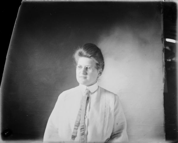 Waist-up portrait in front of a backdrop of Hattie von der Sump sitting. She is wearing a blouse with collar and a necktie.  