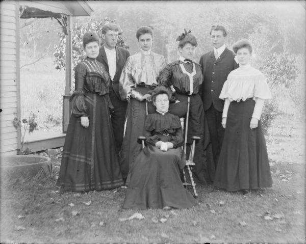 A group of well-dressed young adults posing for a portrait near a porch. They include Margaret (Mae) von der Sump, sitting in the center, and standing behind her, from left, are: Leta and Will von der Sump, Clara ____, Hattie and Fred von der Sump, and Ethel Wallace. The Von der Sumps pictured are siblings. The negative is dated "Oct. 3."