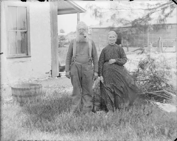 James Cheney and his wife Mary posing outdoors near the side of a house which has a porch. There is a fence and granary in the background, and a tree limb is on the ground at right. There is a wooden half barrel or wash tub on the ground on the left. A dog standing at their feet is blurred from motion. James, who has a full beard, has a pipe in his mouth.  