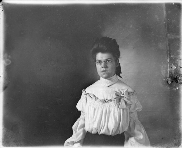 Margaret, also known as Maggie or Mae, von der Sump posing, standing, for a waist-up portrait in front of a backdrop. She is wearing a fancy blouse and has a ribbon in her hair.  