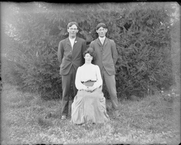 Two young men, identified only as John and Will, are standing behind a young woman, Lilly, who is sitting. Behind the group is an evergreen tree. A man's hat is on the ground at right.