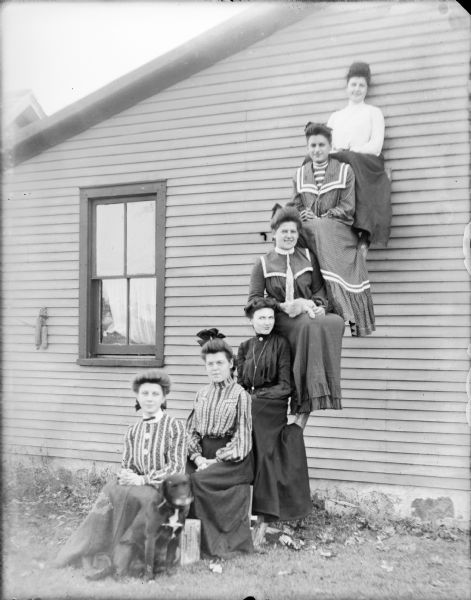 Ethel Wallace is sitting on a wooden box in front of five other women who are sitting on the rungs of a ladder propped on the side of a house. A dog is sitting on the ground beside Ethel. The other women are, from left: Mae von der Sump, Edith Thompson, Hattie von der Sump, Clara _____, and Leta von der Sump at top. Hattie is holding a cat in her lap. The women are all nicely dressed.