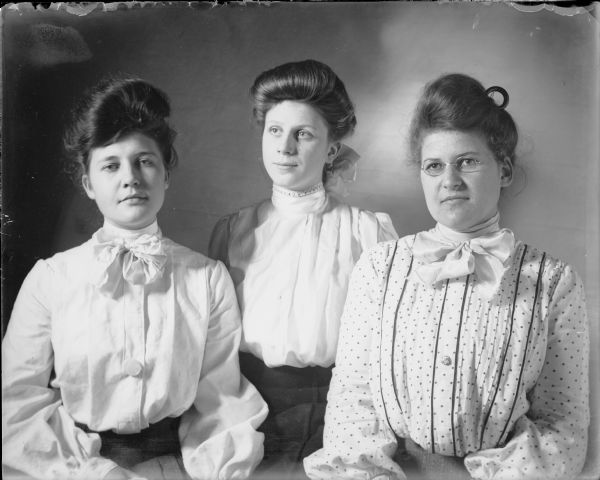 Three  young women, from left Leta von der Sump, Ethel Wallace, and Hattie von der Sump, sitting for a waist-up portrait in front of a backdrop. The women are wearing light-colored blouses with high collars.