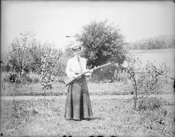 Leta von der Sump posing outdoors holding a rifle. She is wearing a blouse with necktie, a long skirt and a hat. There is a fence and trees behind her.