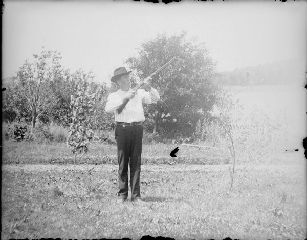 Will von der Sump posing outdoors with a rifle, aiming it upwards. He is well-dressed and is wearing a hat. There is a fence in the background.