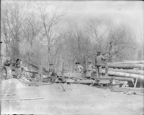 Five men and a boy are posing around a portable sawmill. There is a steam tractor at far left. A large belt is running from the tractor to drive the blade at center. There are piles of sawdust in the foreground, and a stack of logs at right.