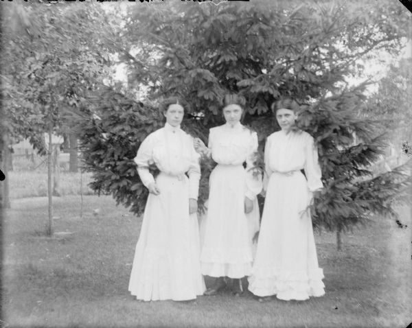Three young women identified only as "Kearns girls" posing outdoors standing in front of an evergreen tree. They are wearing similar long, light-colored dresses with high collars and long sleeves.